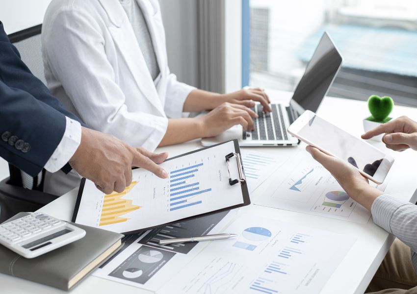 Three property managers discuss a marketing plan for a landlord property, their hands pictured going over paper and online figures and data over an office desk.