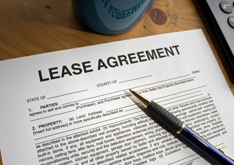 A paper document of a lease agreement sits on a wooden desk with a metal pen on top, waiting to be signed by both the tenant and the landlord.