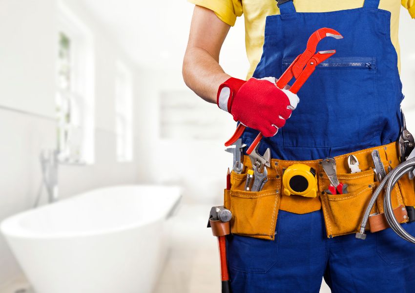 The bottom half of a maintenance worker is pictured with a blue apron, toolbelt, red glove and wrench as they prepare to repair the bathroom plumbing.