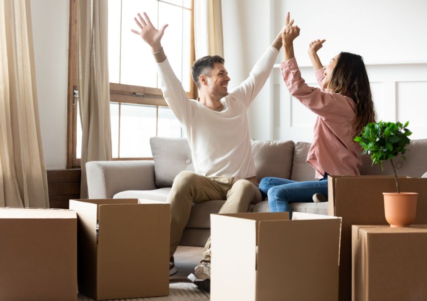 Two smiling tenants, surrounded by move-in boxes in a rental property, raise their arms in the air in excitement.